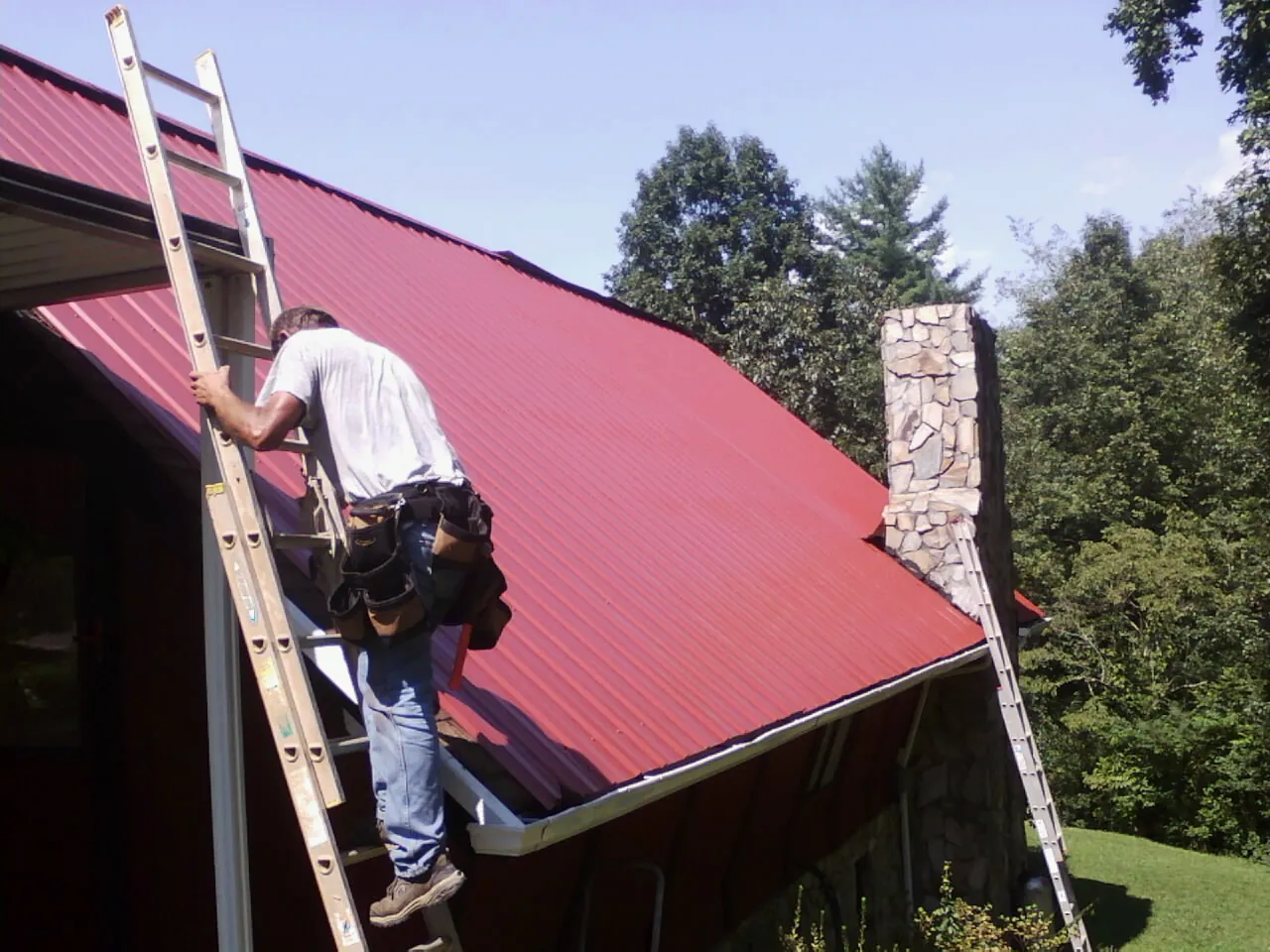 Roofer on a ladder beside a red metal roof and stone chimney — Soffit & Fascia Repair by Roofing Brothers in Macksburg